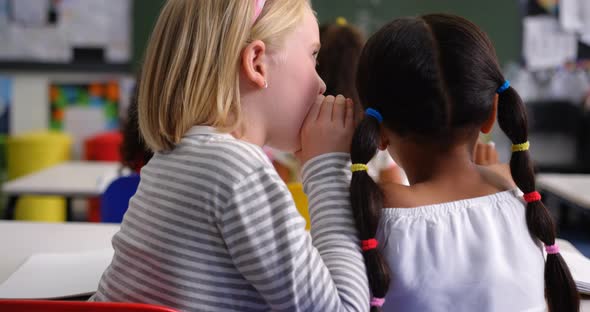 Rear view of mixed-race schoolgirl whispering in her classmates ear at classroom 4k