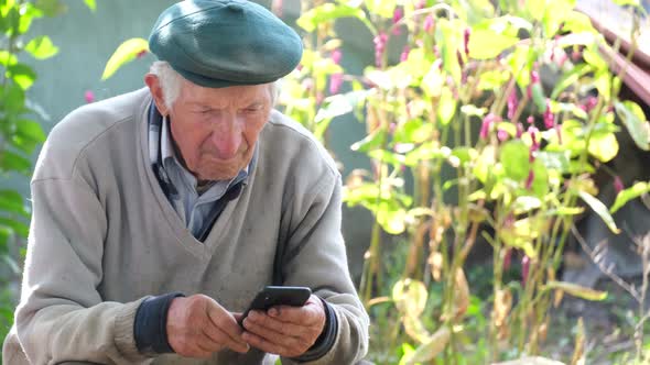 Grayhaired Grandfather Uses a Smartphone While Sitting on a Chair Near the House