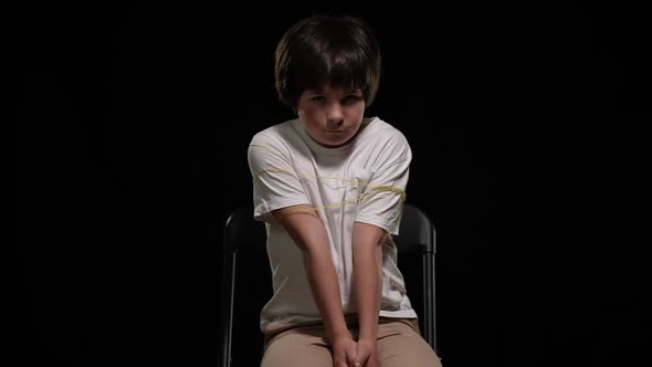 Kidnapped Little Cute Boy with Bruised Face Tyed with Ropes Sitting on Chair Looking at Camera