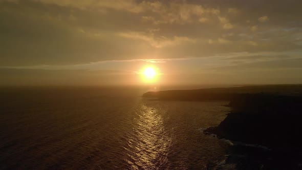 Aerial shot of the orange round sun on the horizon reflecting on the ocean with rugged cliffs in Vic