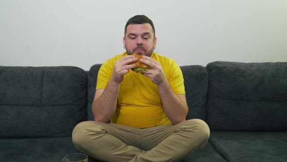 Plump Young Man with Brown Eyes is Sitting on Gray Sofa and Eating Big Fat Burger