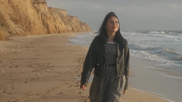 Portrait of Romantic Brunette Woman in Leather Jacket and Dress Walking on the Beach in Autumn at