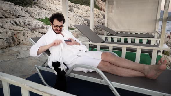 Young Caucasian Bearded Man is Resting in Lounge Area Lying on Sunbed and Playing with a Cat