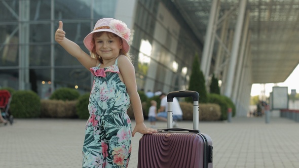 Child Girl Tourist with Suitcase Bag Near Airport. Kid with Luggage Dances, Rejoices, Show Thumbs Up