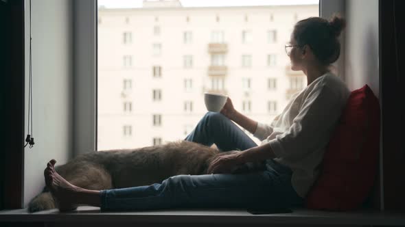 A Young Woman Sits on a Windowsill in Her Apartment with a Dog