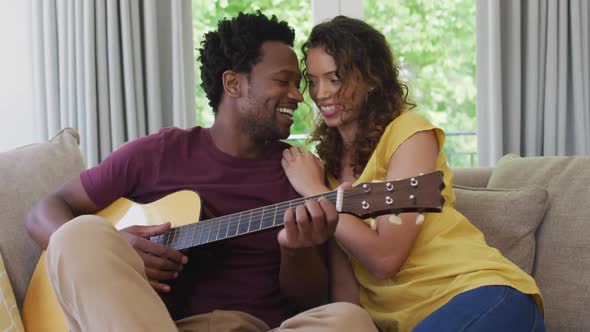 Happy biracial couple sitting on sofa together and playing guitar