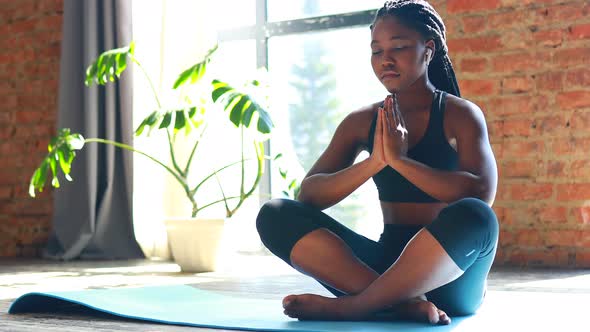 African American Woman in Black Sporty Bra is Sitting Lotus Pose on Yoga Mat Floor Closed Eyes and