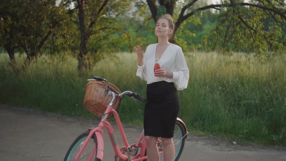 Beautiful Tired Businesslady Standing in Summer Park with Bicycle and Drinking Juice. Portrait of