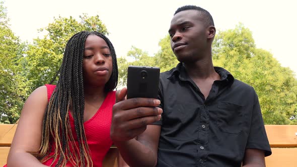 A Black Man and a Black Woman Sit on a Bench in a Park, Look at a Smartphone and Talk