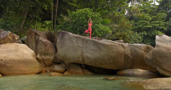 Woman Meditating She Stands on the Big Rock in Pink Sportswear in Yoga Pose and Looks to the Sea