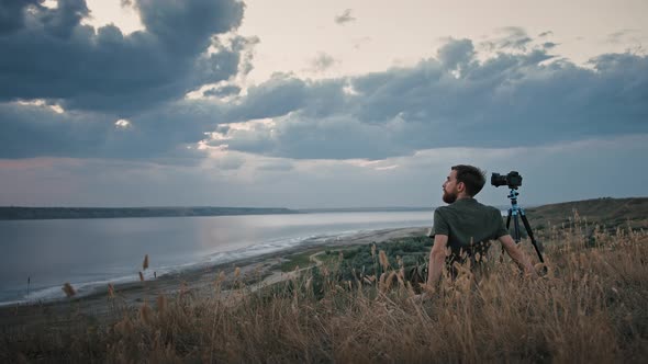 Young Photographer Sitting on High Hill Next to His Camera Fixed on Tripod