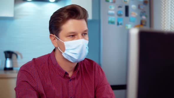 A Man in a Mask Works at Home at a Computer in the Kitchen