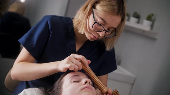 Face Massage  Woman Therapist Using a Big Ribbed Wooden Tool on the Face of Her Female Client