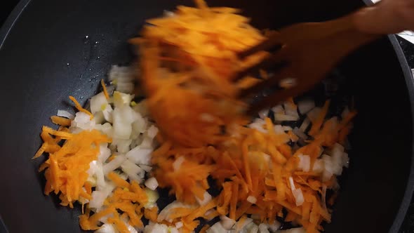 Top view of a woman's hands pouring sliced carrots and onions into a black wok with heated oil