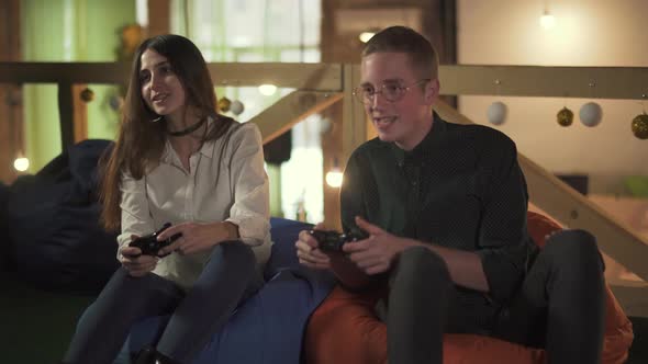 Young Man and Woman Playing Video Game Sitting in Comfortable Chairs in the Cafe. Cute Couple Have a