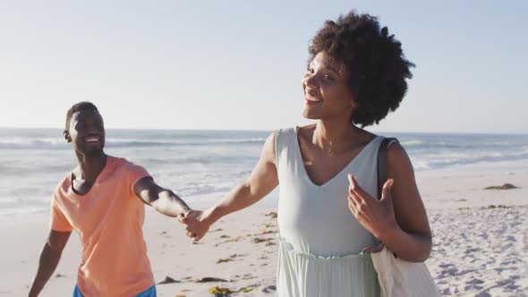 Smiling african american couple holding hands and walking on sunny beach