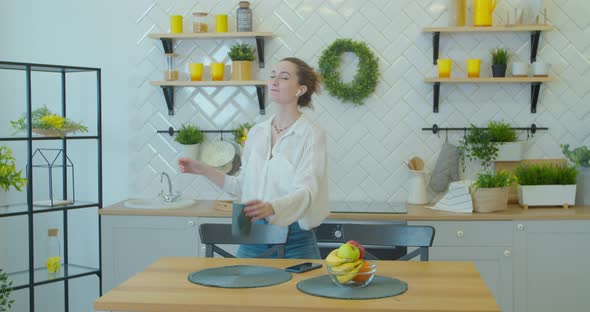 Happy Young Woman Dancing in Kitchen in the Morning Listening To Music on Smartphone and Coffee at