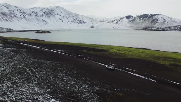 Aerial of a Car Driving on Dirt Road Along Lake and Majestic Mountains Iceland
