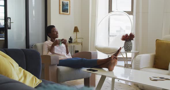 Happy african american woman sitting on armchair in living room, drinking coffee
