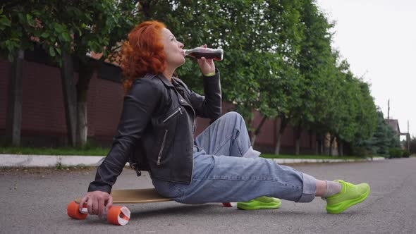 Side View Redhead Retro Woman Drinking Soda Water Sitting on Skateboard Outdoors on Town Street