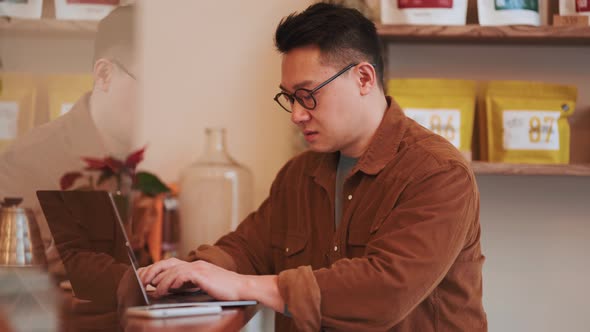 Meditative Asian young man wearing eyeglasses texting by laptop