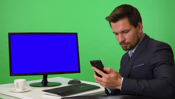 A Young Businessman Sits in Front of a Computer and Works on a Smartphone - Green Screen Studio