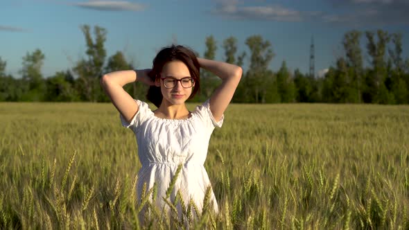 A Young Woman in a White Dress Stands in a Green Wheat Field and Touches Her Hair. Girl Enjoys the