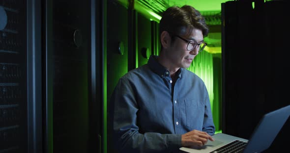 Smiling asian male it technician using laptop checking computer server