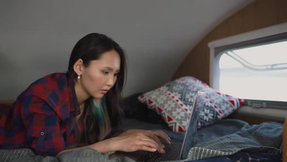 Asian Woman Freelancer Working at a Laptop While Lying in a Mobile Home Typing Text Remote Work in