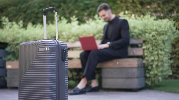 Closeup Travel Bag with Blurred Positive Young Businessman Surfing Internet on Laptop at Background