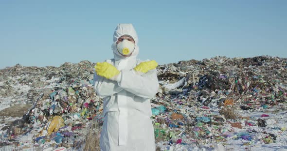 Man in Protective Costume and Mask Shows Sigh Stop with Hands Near Landfill