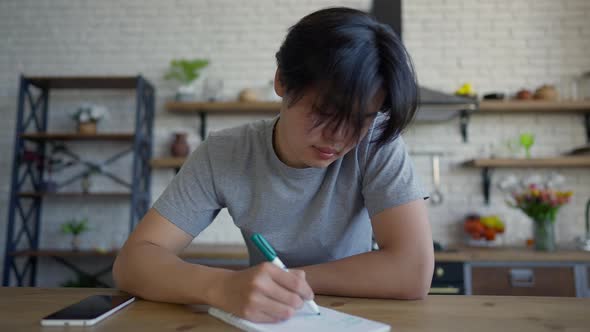 Smiling Concentrated Asian Man Writing with Pen in Sketchpad Sitting at Kitchen Table Indoors