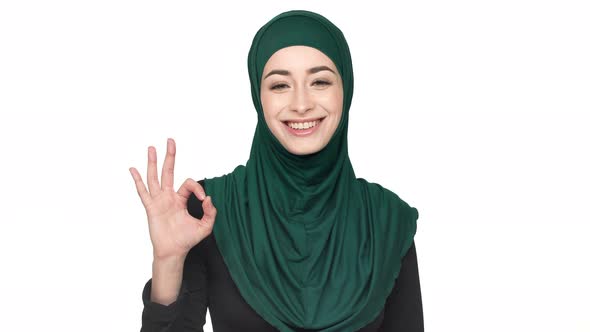 Portrait of Happy Muslim Woman 20s in Traditional Headscarf Showing OK Sign with Fingers and Smiling