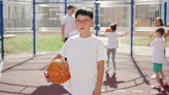 Portrait of Little Asian Boy in Glasses Standing with Ball Outdoors in School Basketball Cours