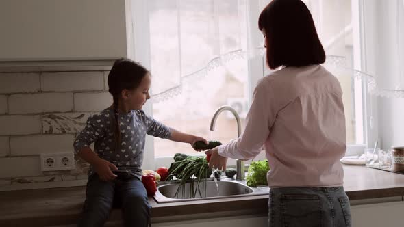 Happy Young Caucasian Mother with Cute Daughter in the Kitchen Wash Vegetables in the Sink