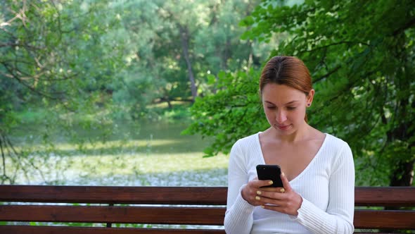 Young Woman Looks at Her Phone and Smiles in the Park.