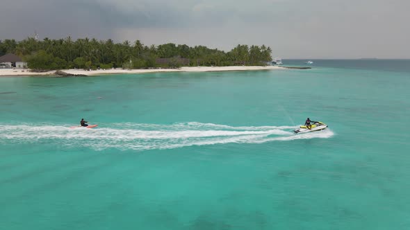 A jet ski goes through the turquoise water next to the island on which green trees grow and the hote