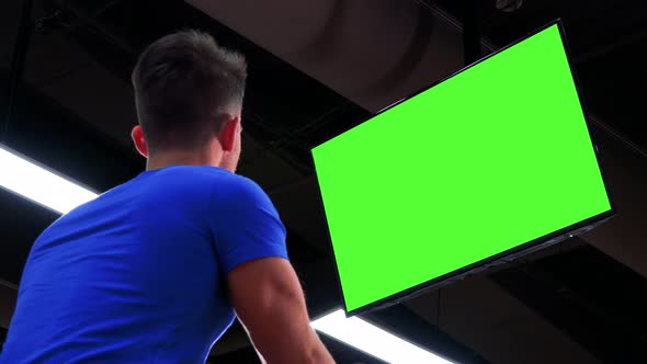 A Young Fit Man Trains on an Exercise Bike in a Gym and Watches a Green Screen - View From Below