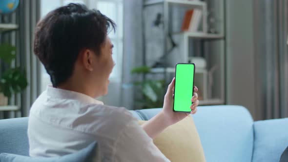 Man Waving Hand And Talking To Green Screen Smartphone While Lying On Sofa In The Living Room