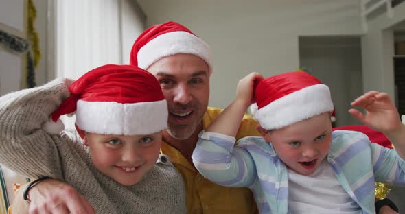 Portrait of caucasian father and two sons wearing santa hats waving and smiling sitting on the couch