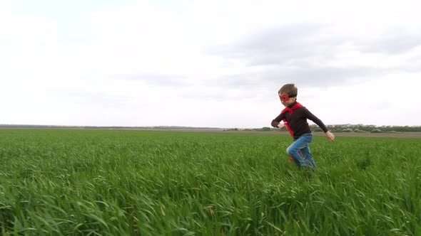 A Happy Child in a Superhero Costume in a Red Raincoat Runs Across a Green Lawn Against a Blue Sky.