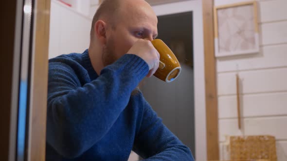 A Man Sits at Home in the Evening at the Table Drinking Coffee in Front of a Laptop Screen