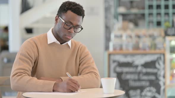 African American Man Writing on Paper While Drinking Coffee in Cafe