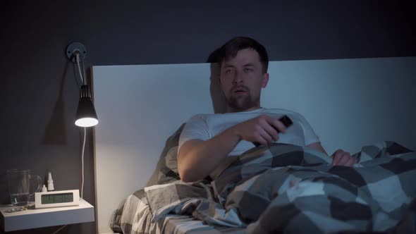 Tired Man Holds a TV Remote Control in Hands and Falling Asleep Watches TV at Night in Bed