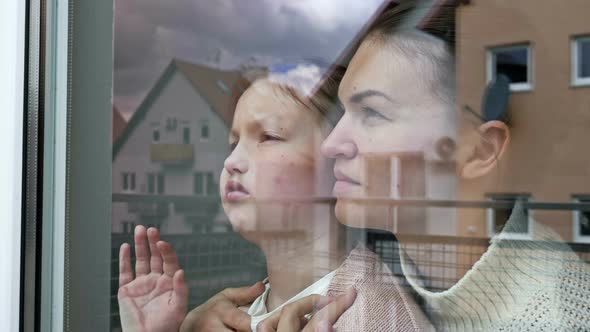 Mother and Little Daughter Stand Embracing and Look Sadly Out the Window