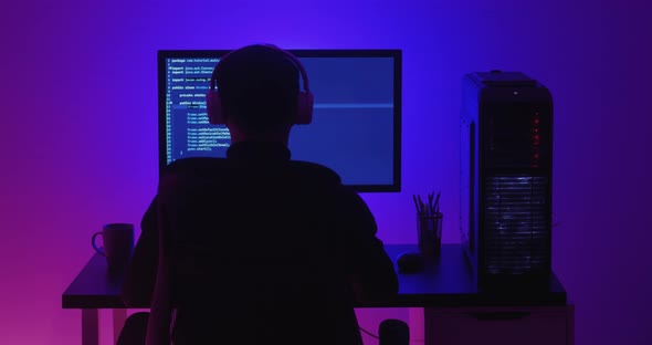 Rear View of Man Programmer in Headphones Coding Network Security Software Indoors in Neon Lights at