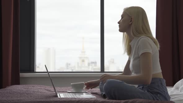 Beautiful Young Woman Works Behind a Laptop in the Bedroom with a Large Window