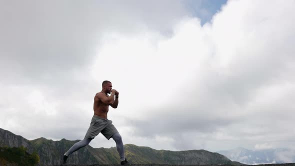 a Man with a Bare Muscular Torso is Boxing Against the Background of Mountains and Low Clouds