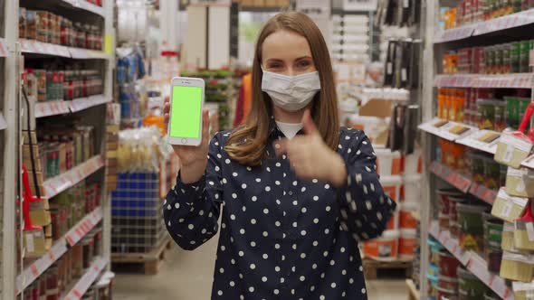 Woman Wearing a Protective Mask Pointing at the Green Screen of a Smartphone Looks at the Camera in