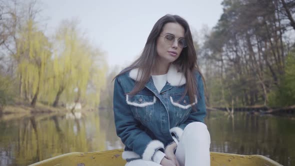 Portrait of a Gorgeous Young Woman in Sunglasses and a Denim Jacket Floating on a Boat on a Lake